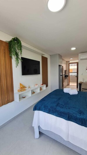 A modern bedroom with a bed, a mounted TV, a small shelf with decorative items, and a view into the adjacent kitchen area.