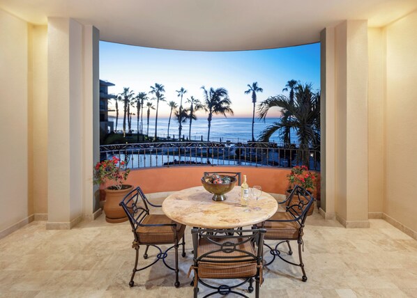Een ronde tafel met vier stoelen, een decoratieve schaal en twee glazen op een balkon met uitzicht op een strand met palmbomen en een hek.