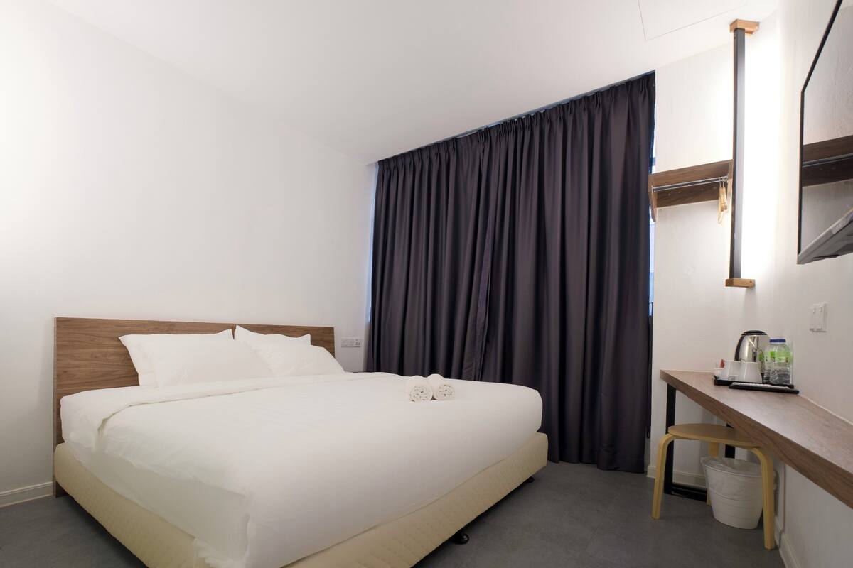 A neatly made bed with white linens, a wooden headboard, a small wooden desk with a chair, a mirror, and a window with dark curtains.