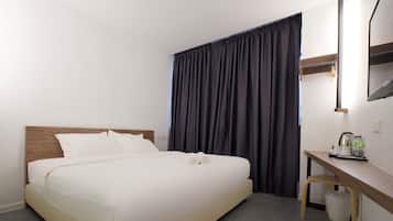 Una cama bien tendida con sábanas blancas, un cabecero de madera, un pequeño escritorio de madera con una silla, un espejo y una ventana con cortinas oscuras.