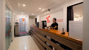 A reception area with a wooden counter, a person behind it, and a wall with a large red logo. - T Hotel Jalan Sultanah (Alor Setar)