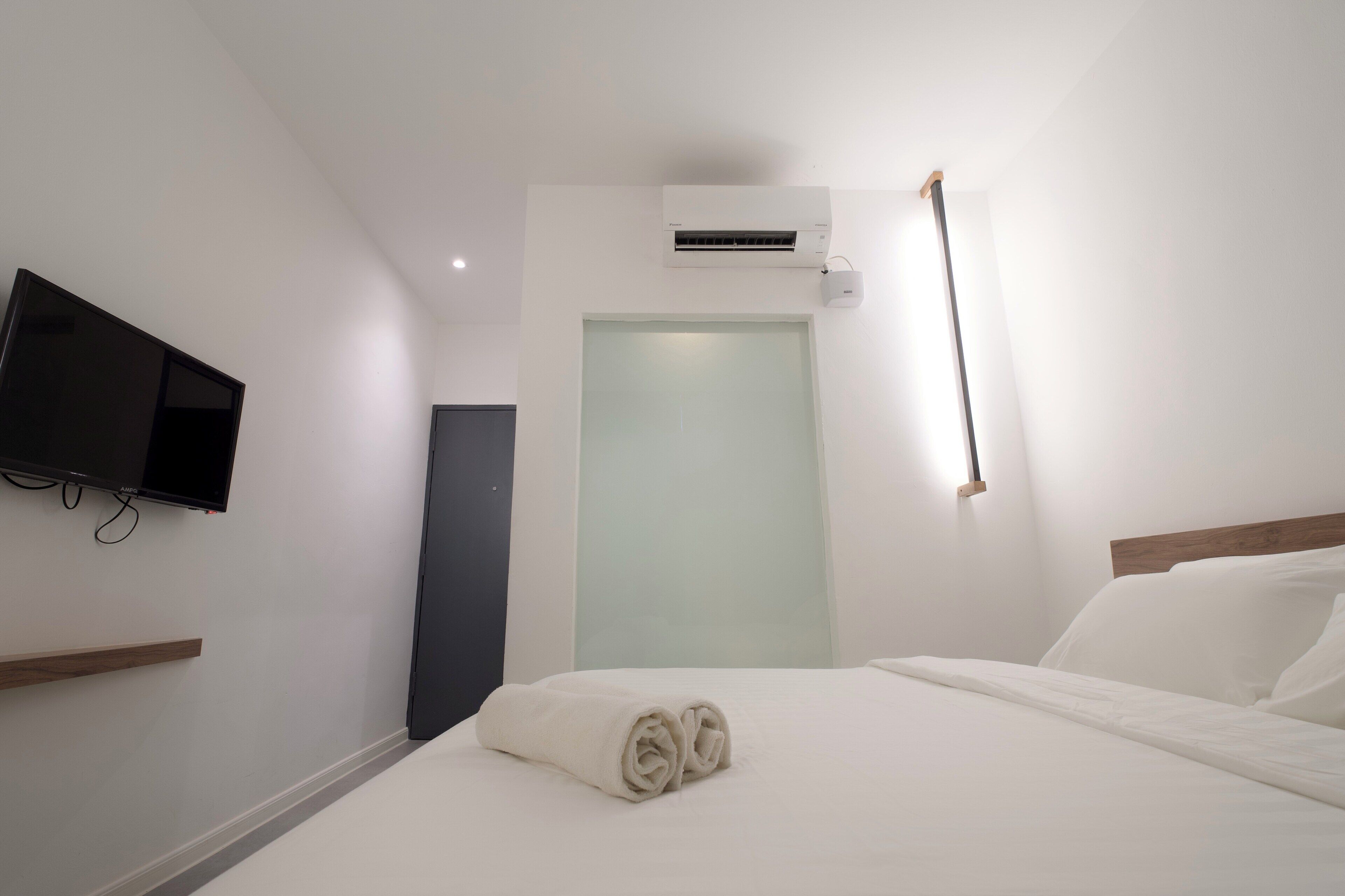 A hotel room with a bed, a flat-screen TV mounted on the wall, an air conditioning unit, and a door with frosted glass.