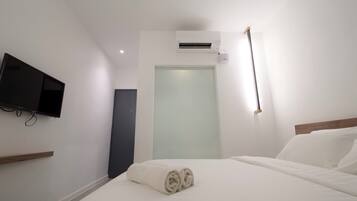 A hotel room with a bed, a flat-screen TV mounted on the wall, an air conditioning unit, and a door with frosted glass.