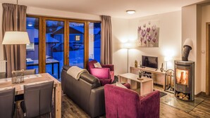 A cozy living room with a sofa, chairs, a table, a TV, a fireplace, and a view of a snowy landscape through large windows. - Residence Lipno by Interhome (Lipno nad Vltavou)
