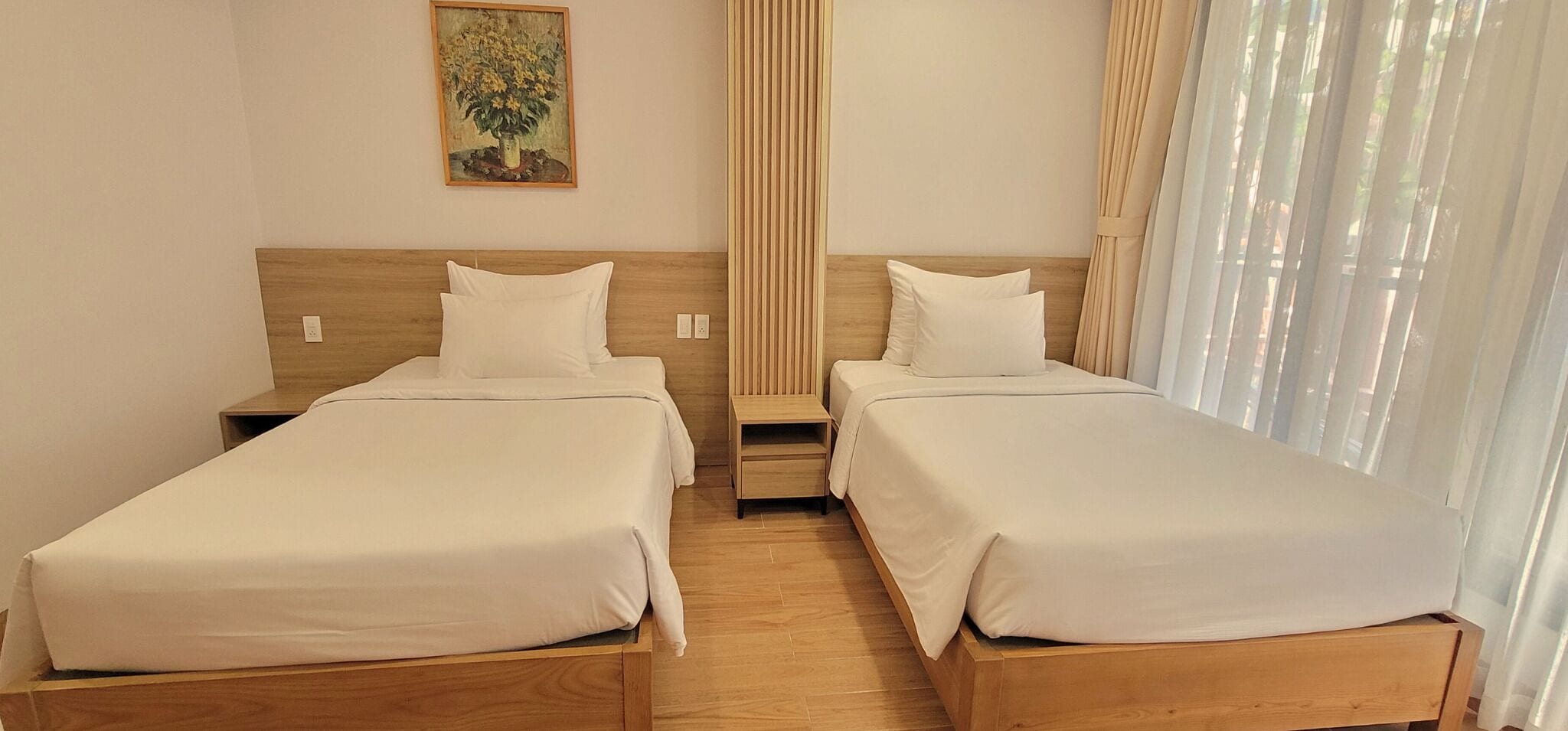 A hotel room with two single beds, wooden headboards, a small bedside table, a nightstand, a painting on the wall, and a window with sheer curtains.