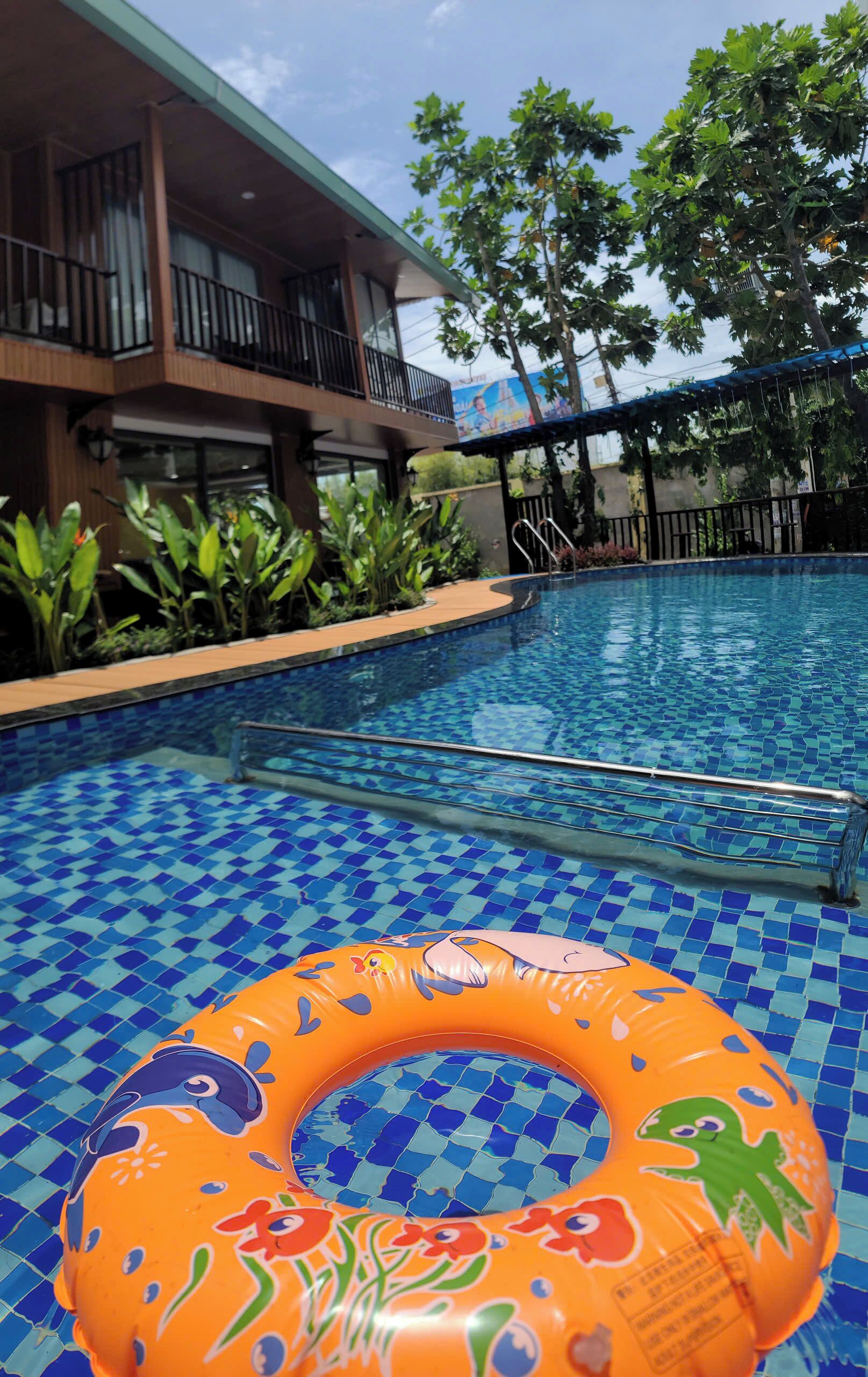 A swimming pool with a tiled floor, surrounded by plants and a building with balconies.