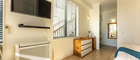 A bedroom with a bed, a TV mounted on the wall, a heater, a window with blinds, and a staircase leading to another room.