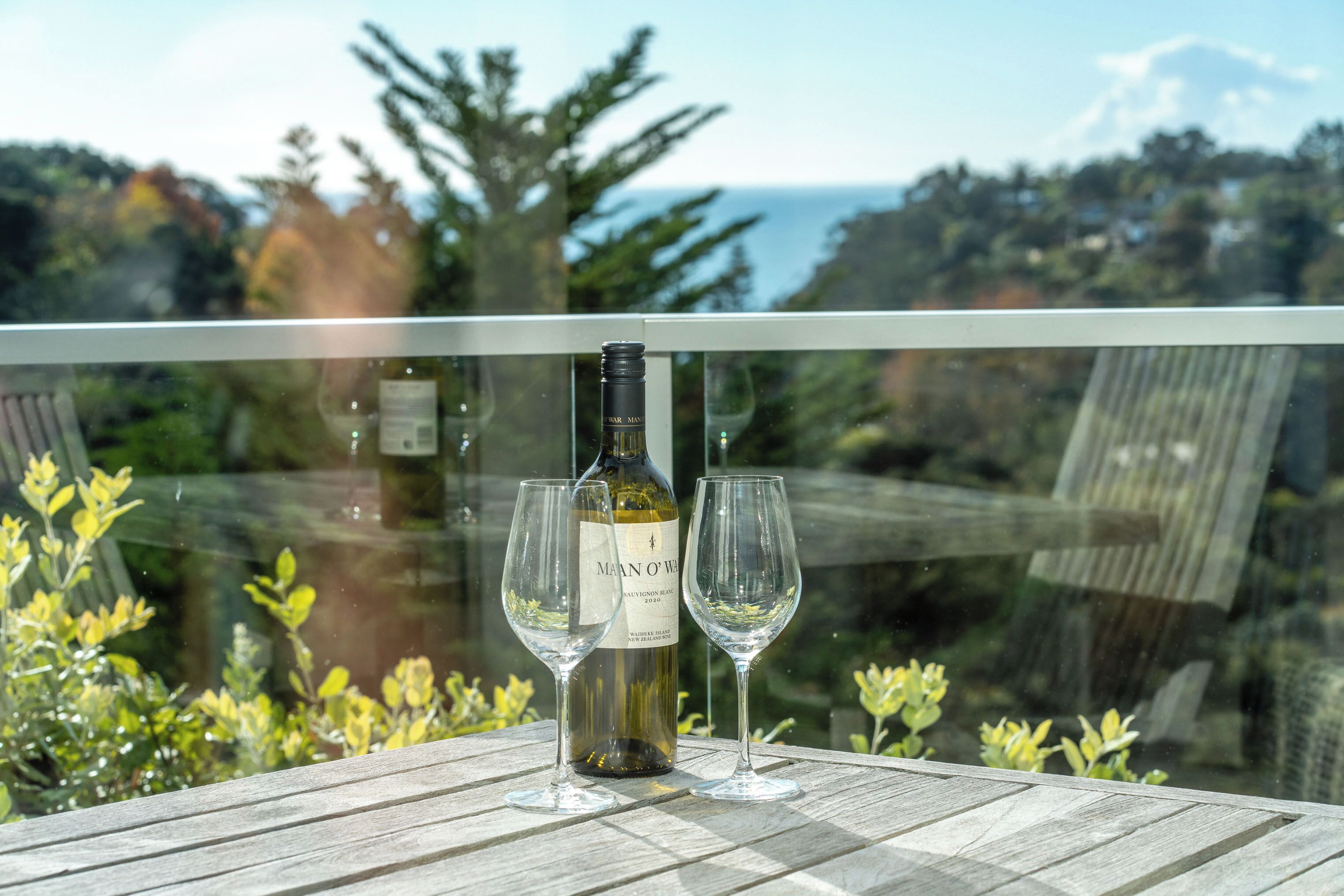 A bottle of Man O' War wine and two glasses on a wooden table with a view of the ocean and trees.