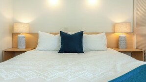Una cama bien hecha con almohadas blancas y azul oscuro, dos lámparas fijadas a la pared y una lámpara con textura en cada mesita de noche.