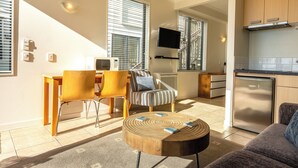Una sala moderna con sofá, una mesa redonda y un comedor con sillas y una mesa. Se observa también una cocina con armarios y un refrigerador.