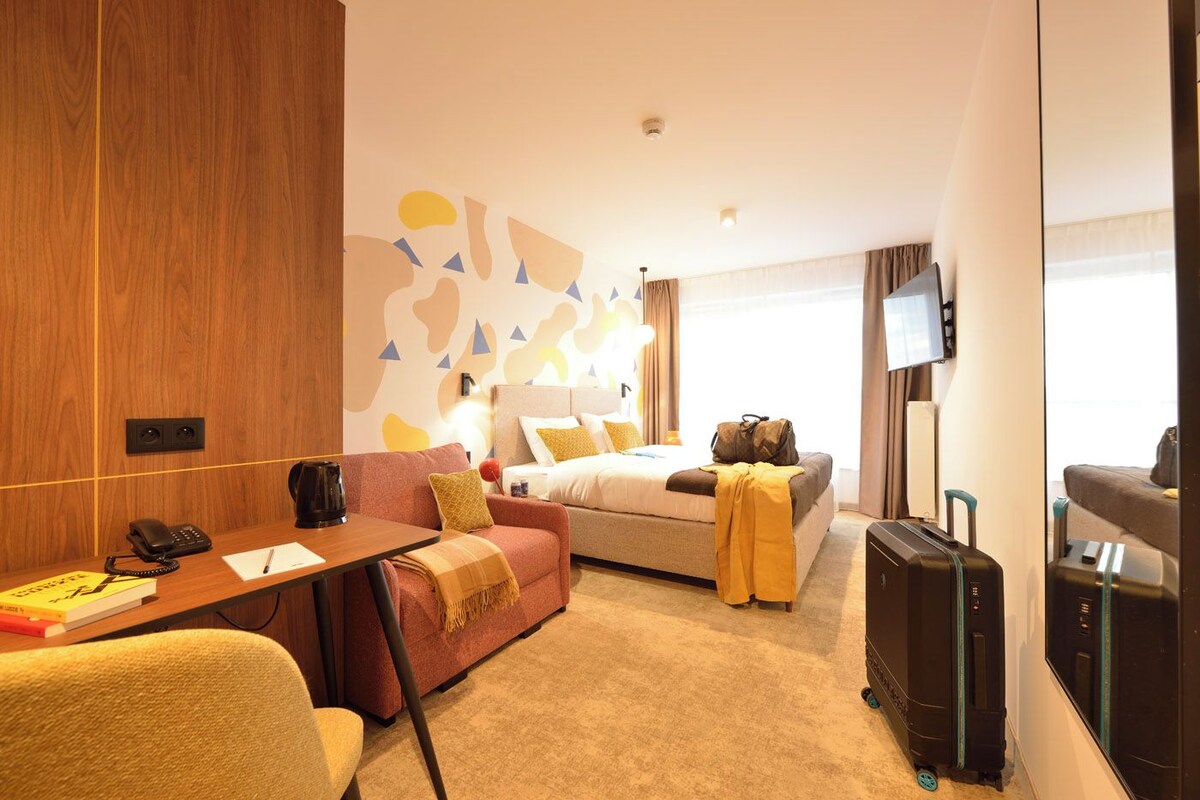 A hotel room with a wooden accent wall, a bed with a beige and brown blanket, a desk with a phone and papers, a sofa with cushions, a TV on the wall, and a suitcase on the floor.