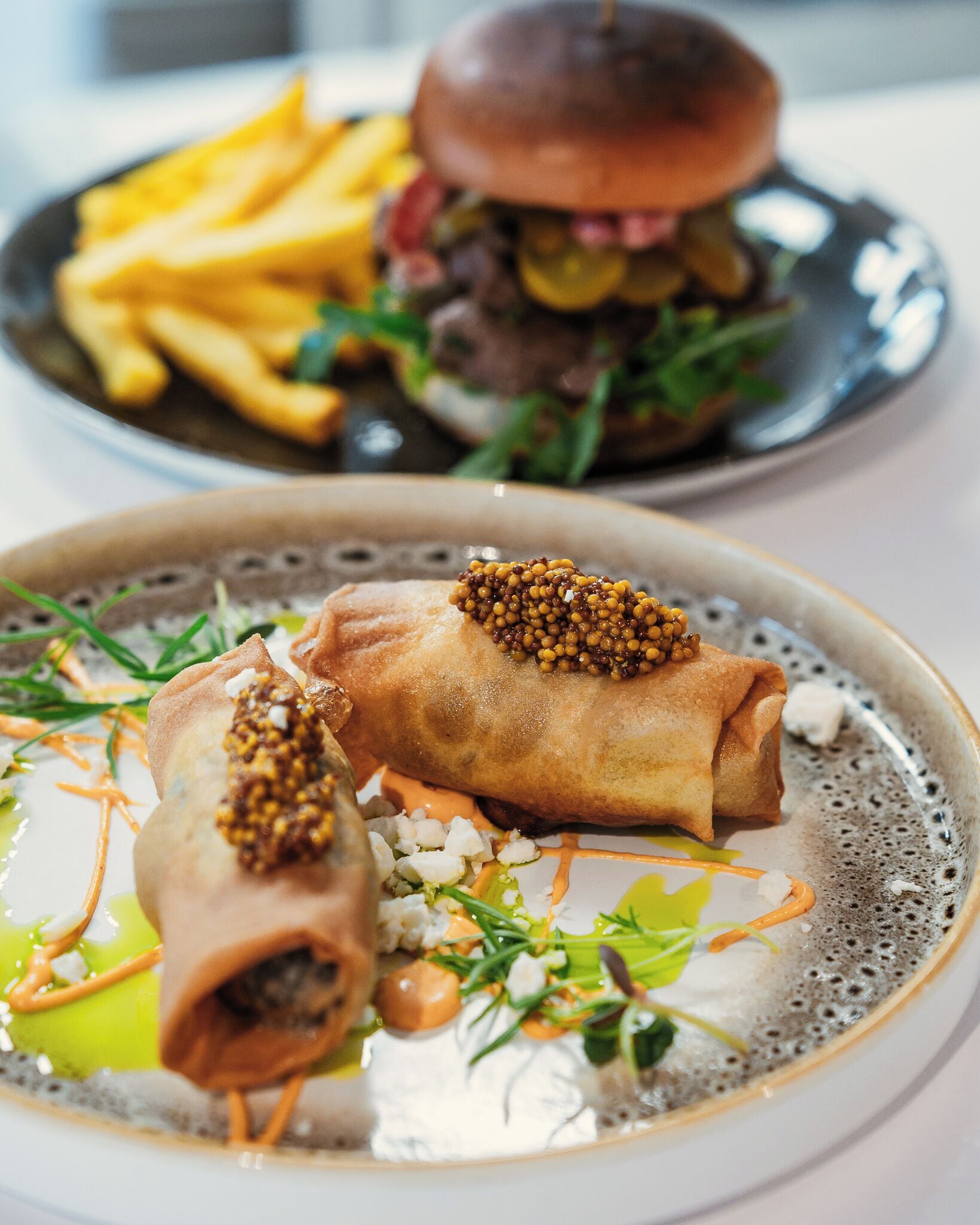 A plate of spring rolls with a sauce and garnish, and a burger with fries in the background.