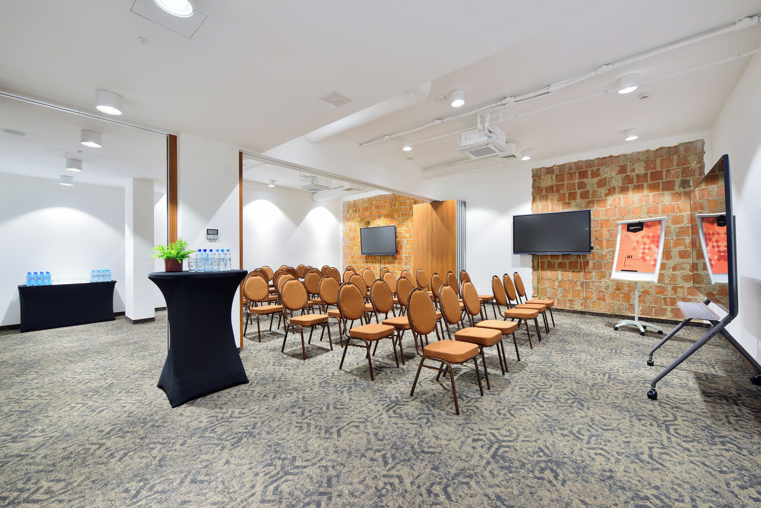 Ein Konferenzraum mit Stuhlreihen, einem schwarzen Tisch mit Pflanze, einer Ziegelwand, einem Fernseher und einer Whiteboard-Leinwand.