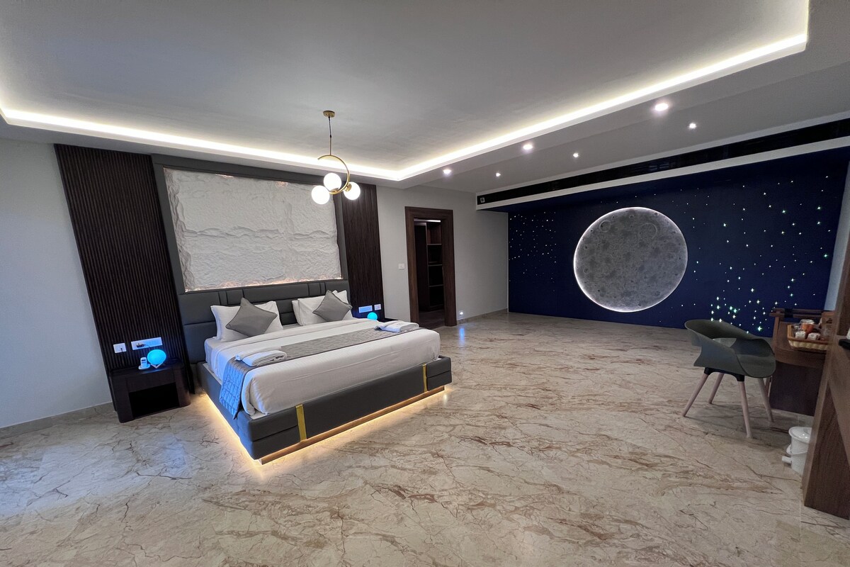 A modern bedroom with a large bed, a nightstand, a desk, and a wall mural of a moon.