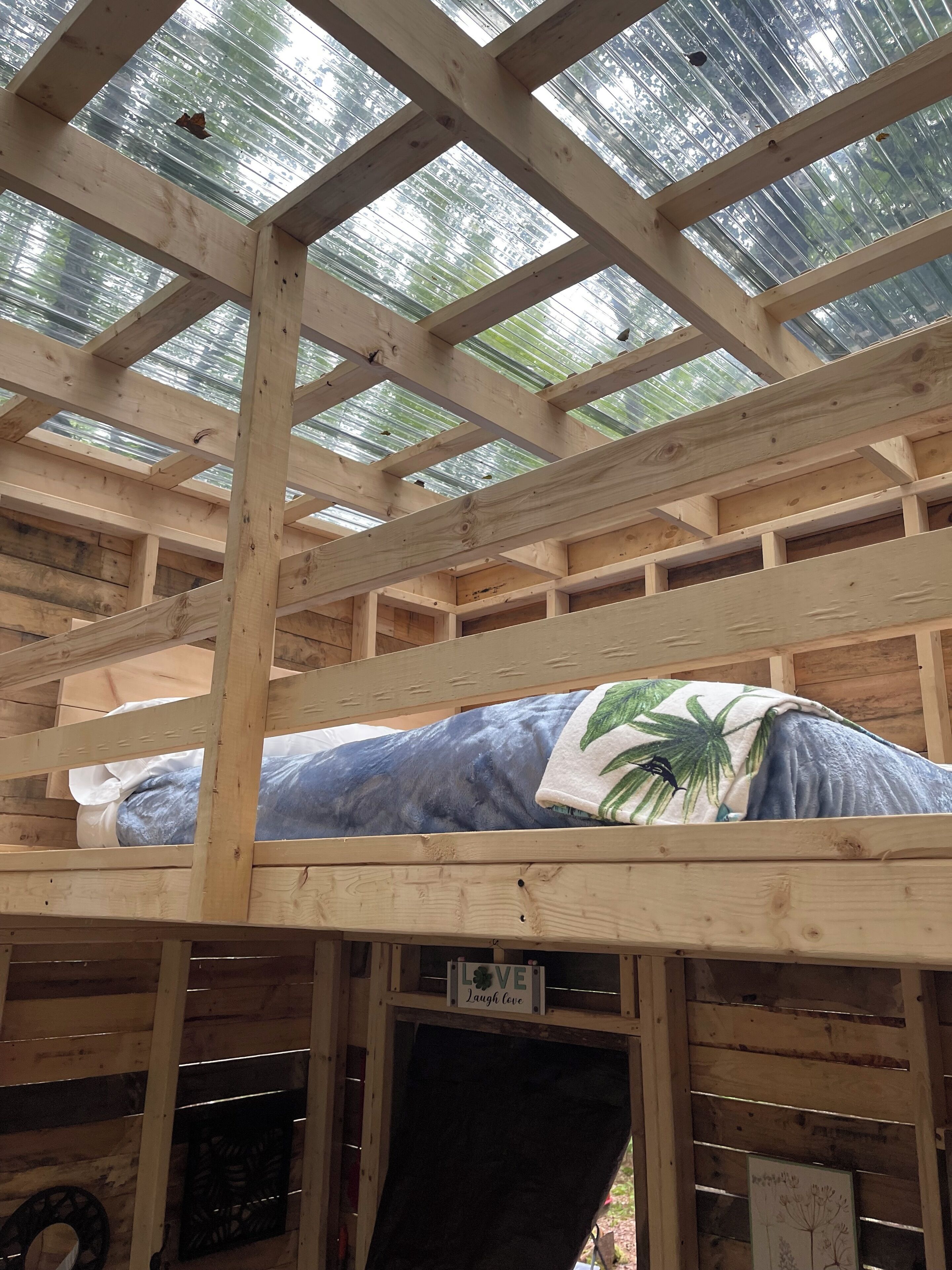 A wooden bunk bed with a mattress covered in a blue blanket with a palm tree design, under a metal roof structure.