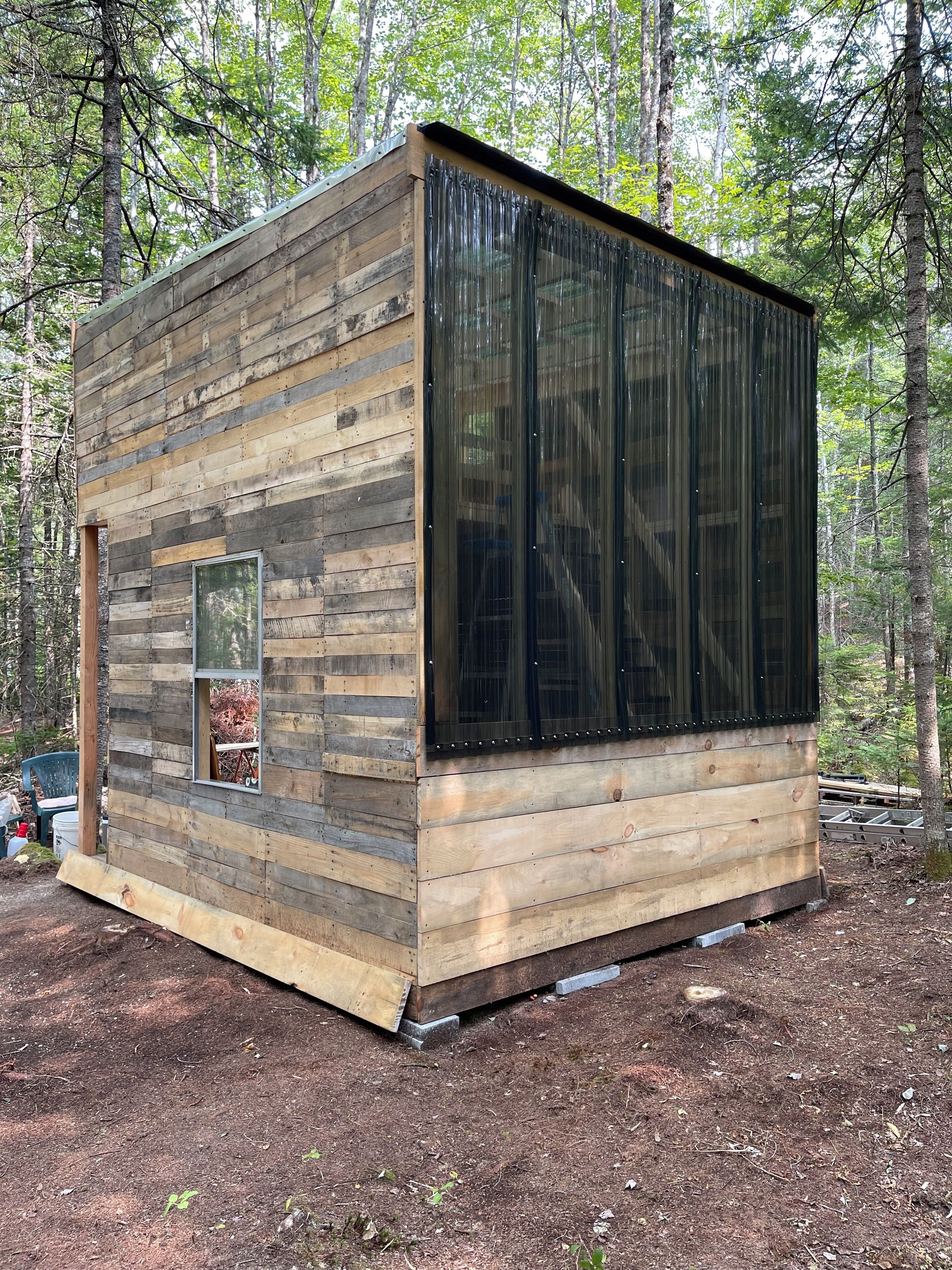 A small wooden cabin with a large window and a door, surrounded by trees.