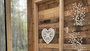 Ein Interieur einer Holzhütte mit einem Bett, einer herzförmigen Wanddekoration und einem Fenster mit Blick auf Bäume.