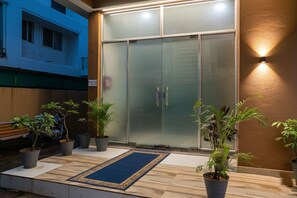 Une entrée d’immeuble avec une porte en verre, des plantes en pot et un paillasson bleu.
