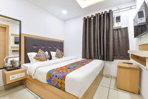 A modern hotel room with a neatly made bed, a small bedside table with a kettle, a power outlet, and a TV on a wooden stand. - Fabexpress Perfect (Samba)