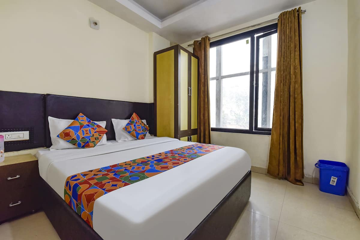 A hotel room with a bed, a nightstand, a window with curtains, and a blue trash bin.