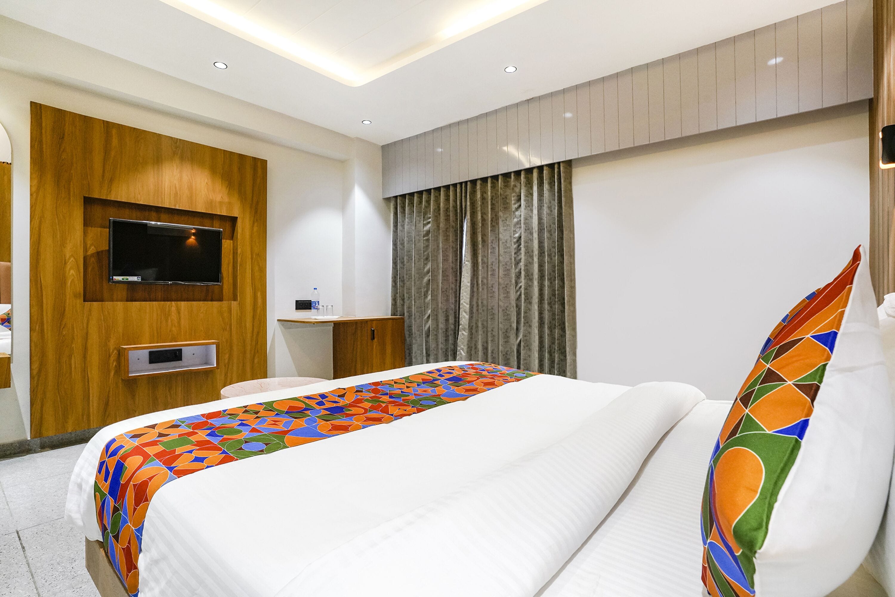 Ein Hotelzimmer mit einem Bett, einem an einer Holzvertäfelung befestigten Fernseher, einem Schreibtisch und einem Fenster mit Vorhängen.