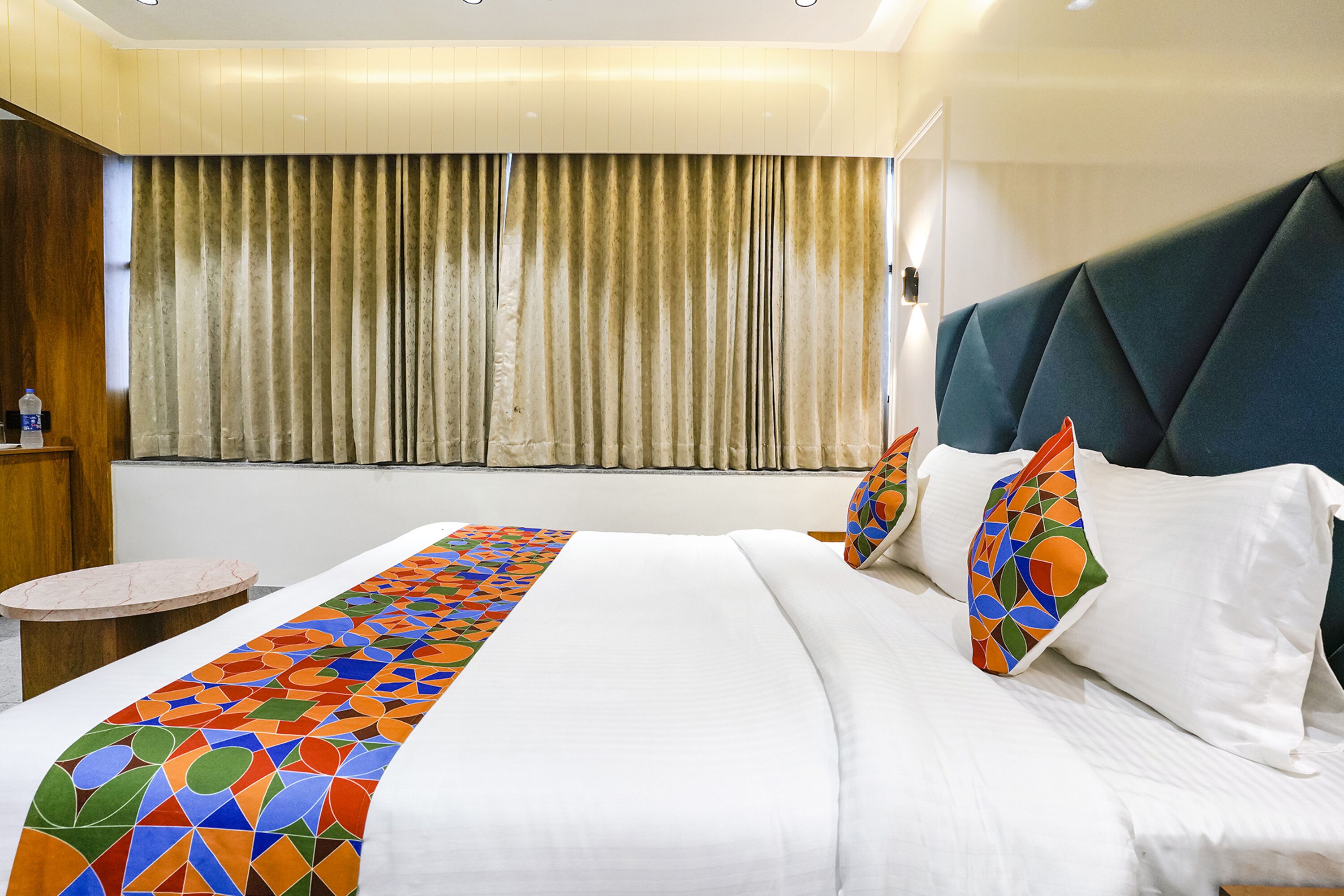 A hotel room with a large bed, a headboard, a small round table, and a window with curtains.