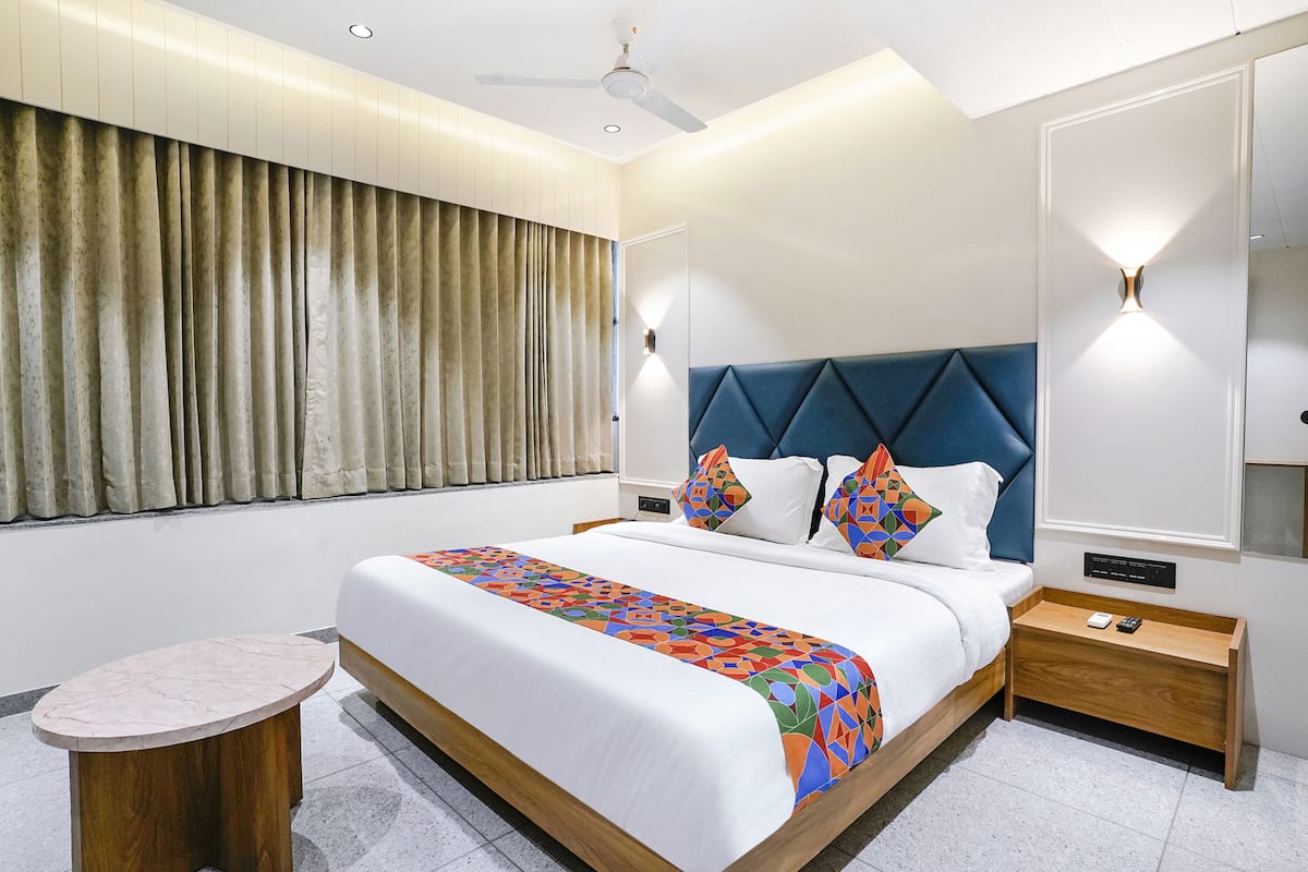 Une chambre d’hôtel moderne avec un grand lit, une petite table d’appoint ronde en bois, un ventilateur de plafond et une grande fenêtre avec des rideaux.
