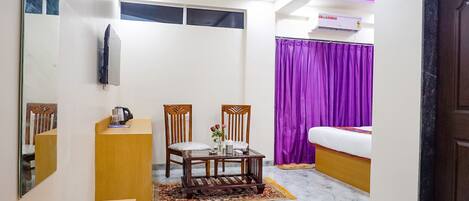 Quarto de hotel com cama, escrivaninha, duas cadeiras, cafeteira, pequena mesa com flores, ventilador de teto, ar condicionado e cortina roxa.