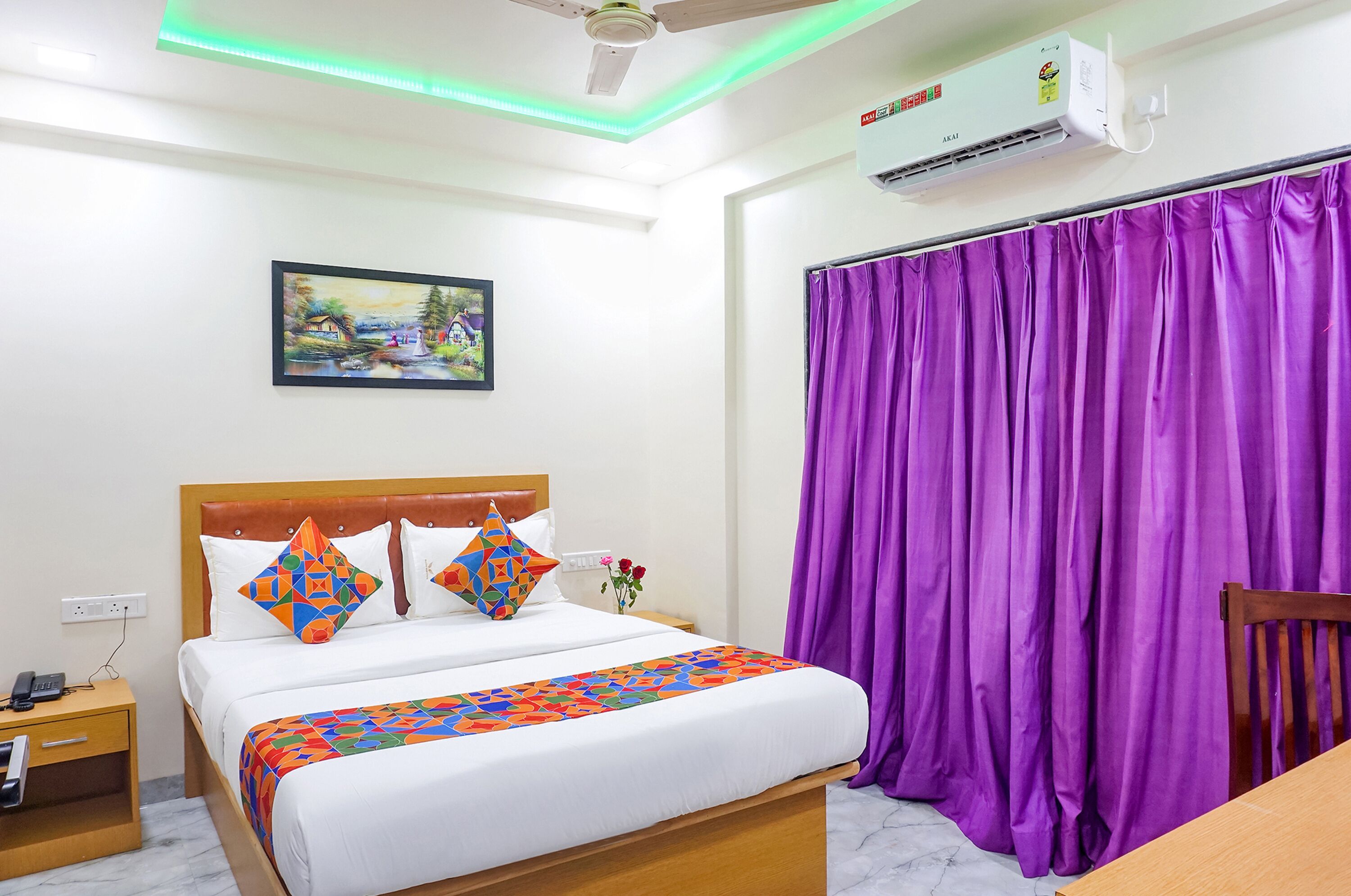 Ein Hotelzimmer mit einem Bett, einem Nachttisch, einer Klimaanlage, einem Deckenventilator, einem violetten Vorhang und einem Bild an der Wand.