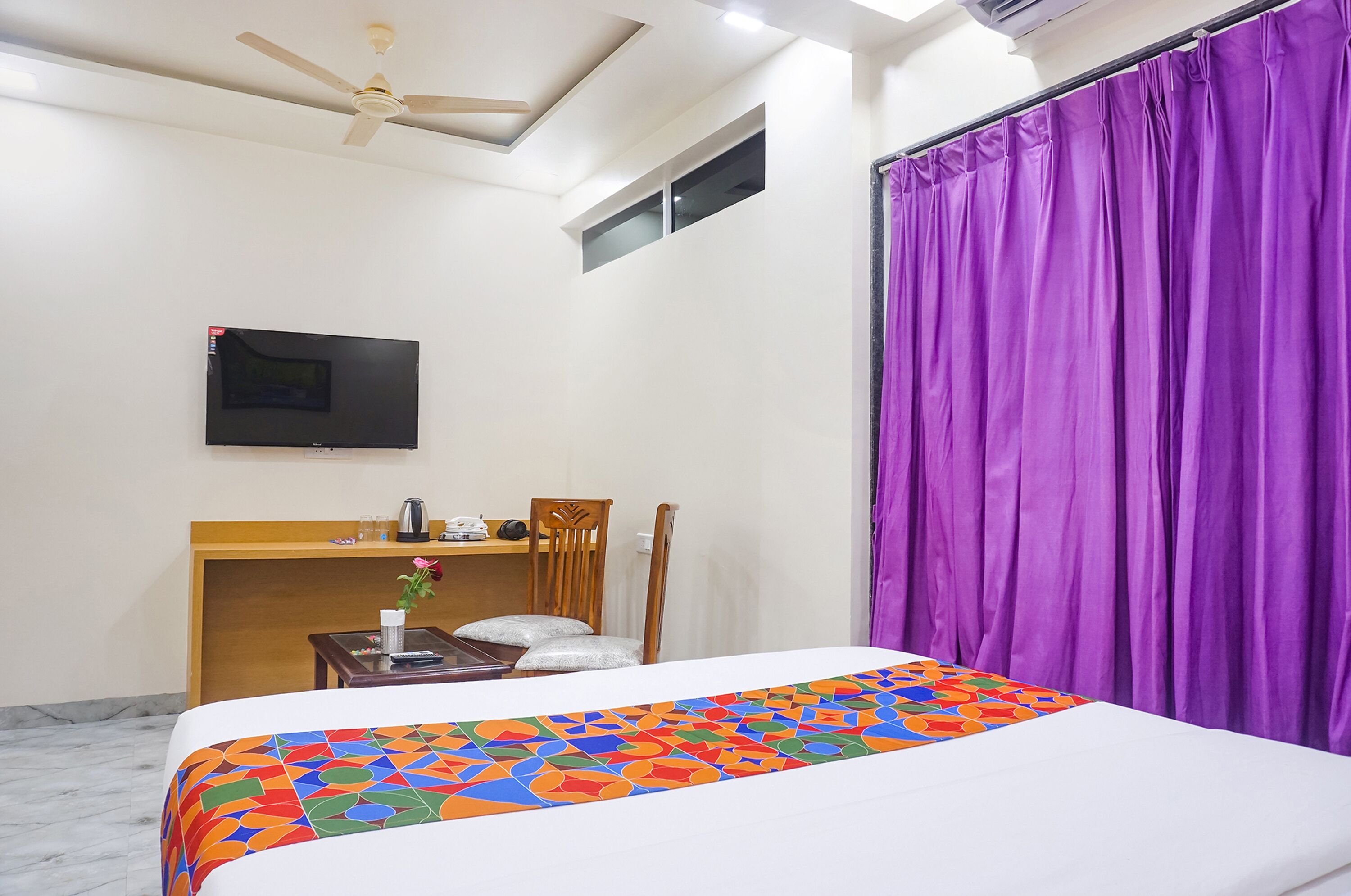 A hotel room with a bed, a desk, a chair, a TV, and purple curtains.
