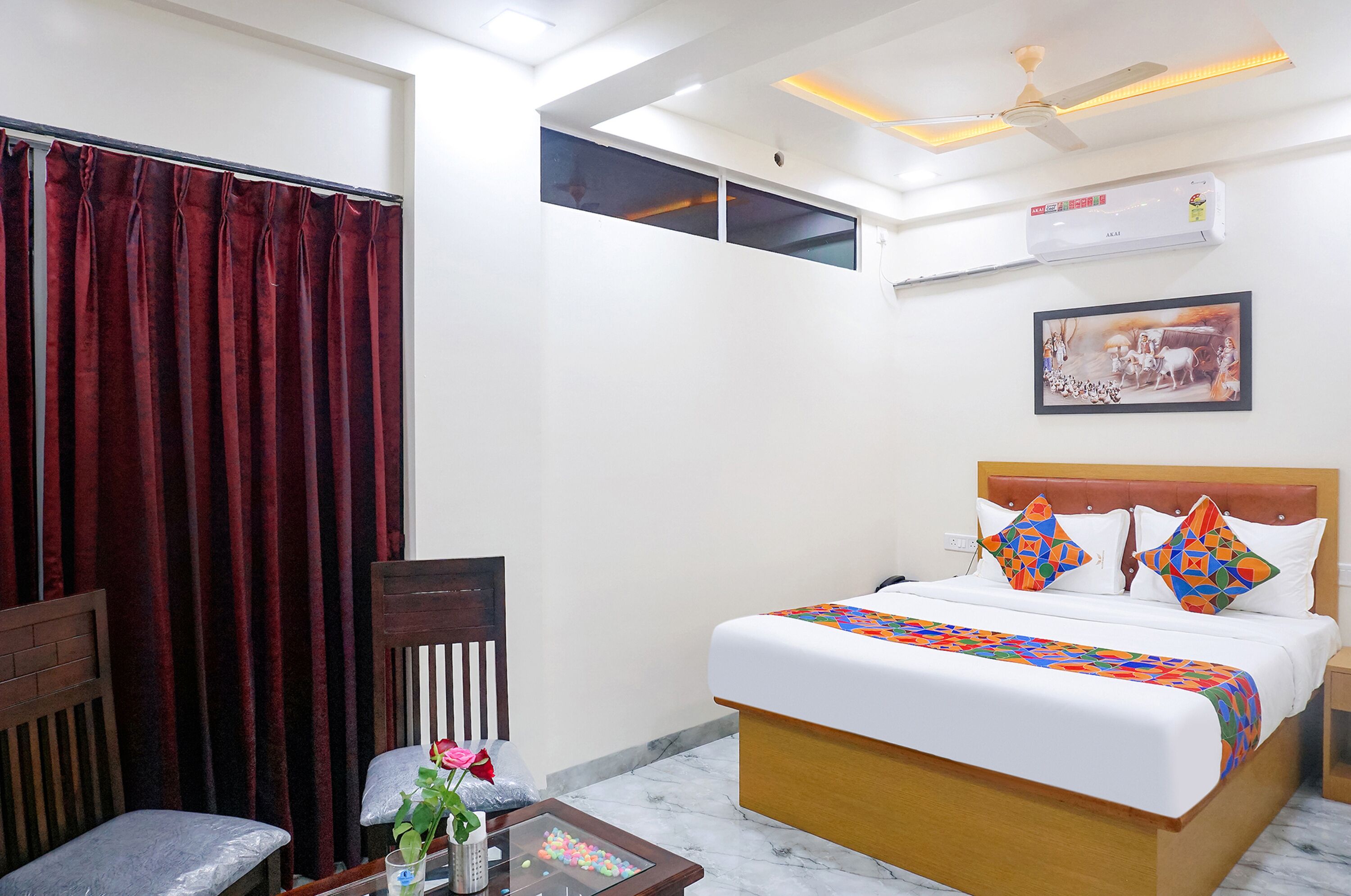 A hotel room with a bed, a chair, a small table with a vase and decorative items, a window with curtains, an air conditioning unit, and a ceiling fan.