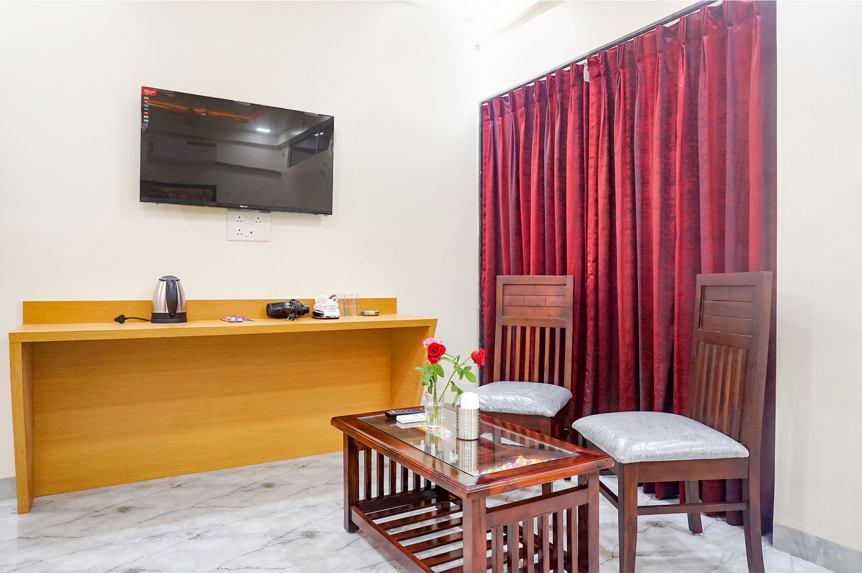 A hotel reception area with a wooden counter, a television mounted on the wall, a red curtain, a wooden chair, and a small table with a vase of flowers.