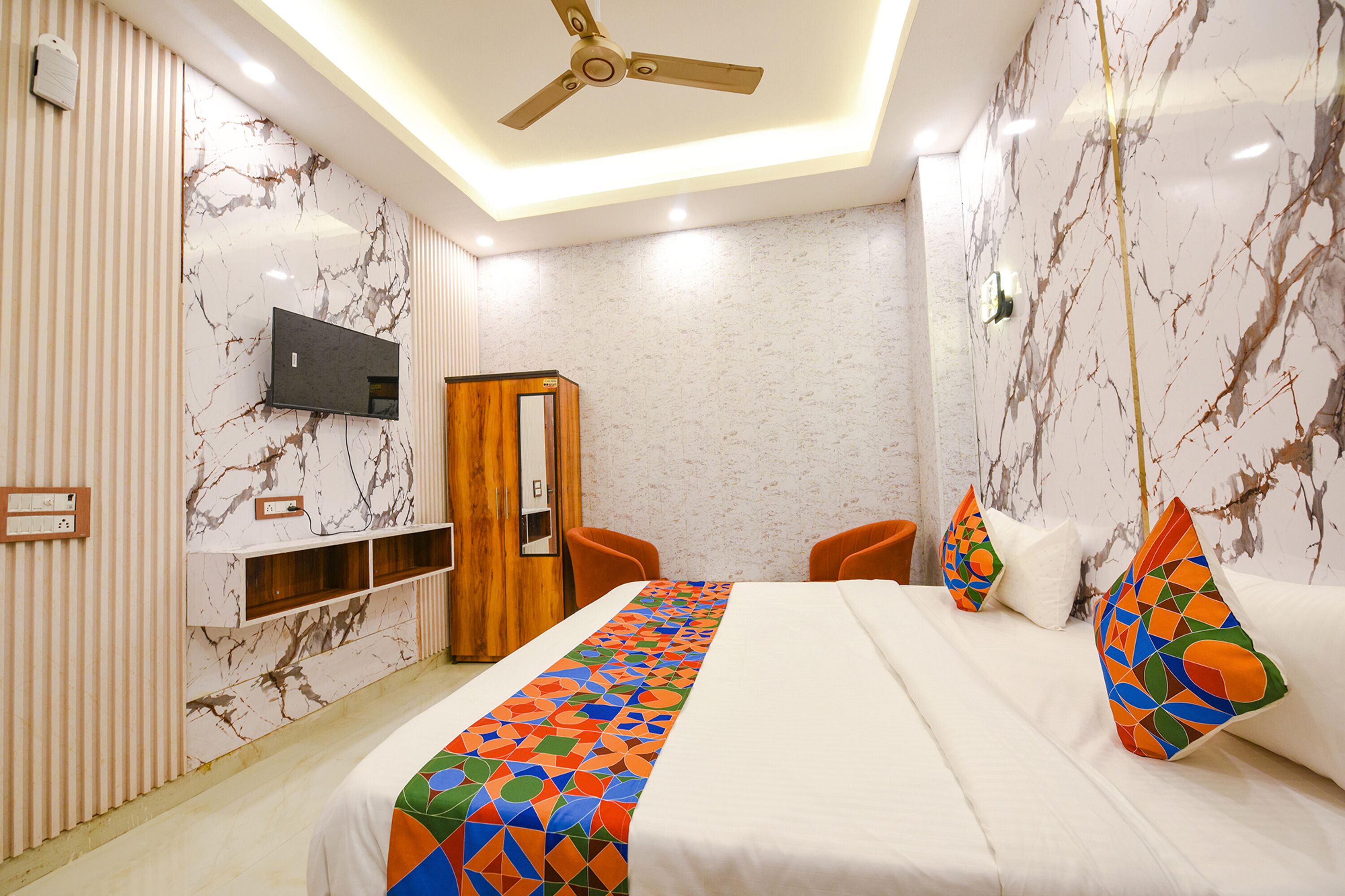 A hotel room with a bed, a TV mounted on the wall, a wooden wardrobe, and a ceiling fan.
