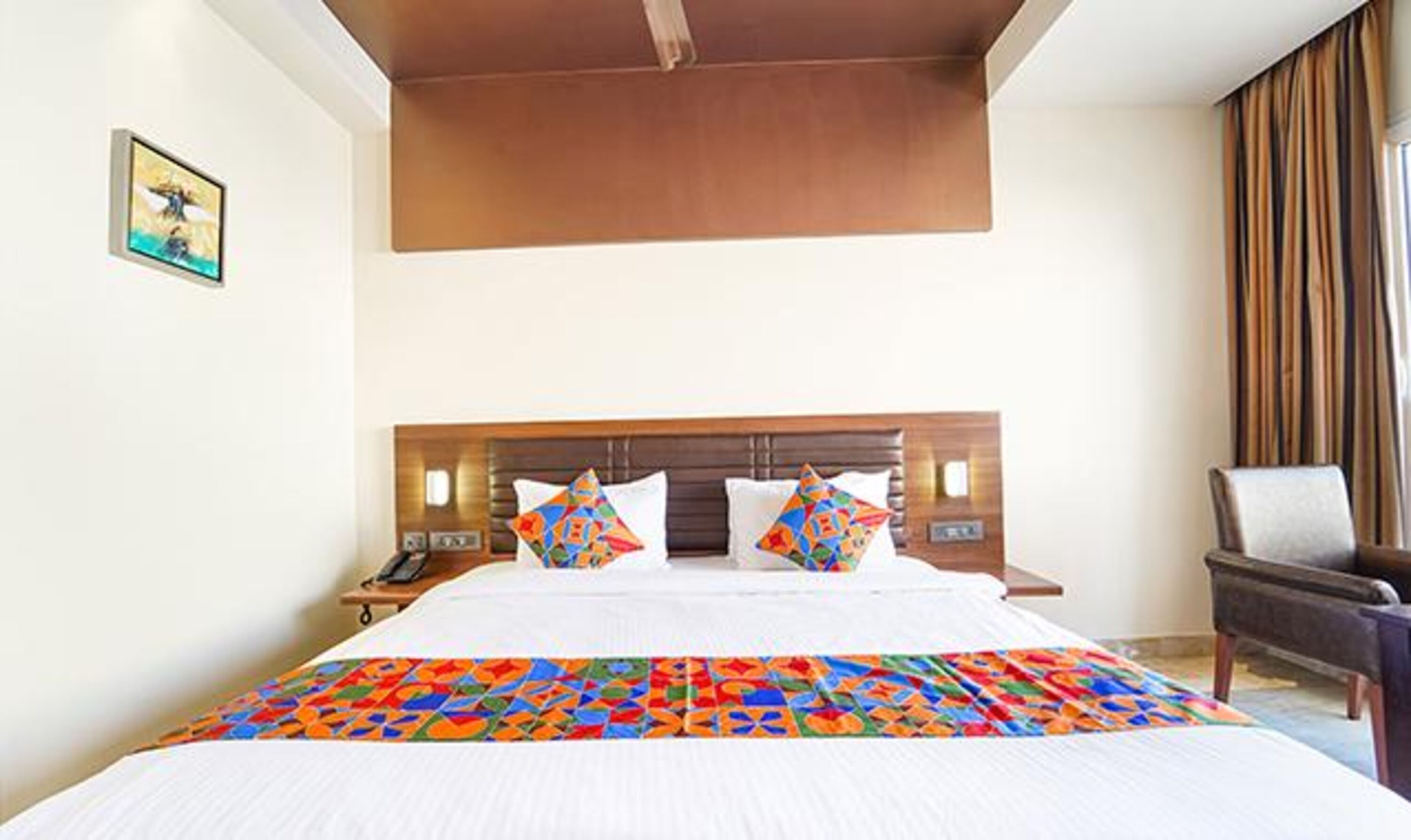 A hotel room with a large bed, a wooden headboard, a nightstand with a phone, a chair, and a painting on the wall.