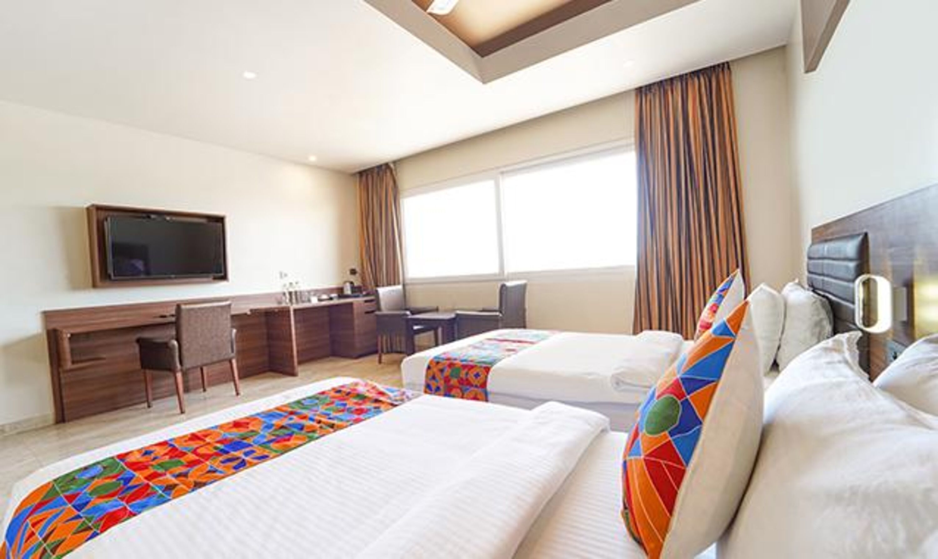 A hotel room with two beds, a desk, a TV, and a large window with curtains.