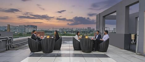 A rooftop terrace with a checkered floor, outdoor seating, and a cityscape view at dusk.