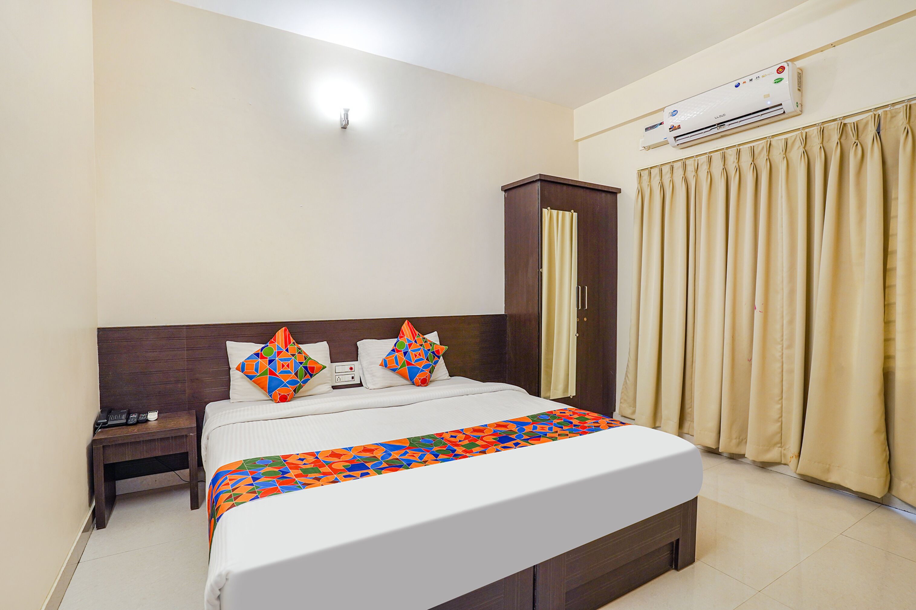 A hotel room with a bed, a nightstand, an air conditioning unit, and a window with curtains.