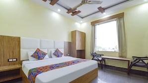 A hotel room with a large bed, wooden headboard, a bench, and a window with curtains. - Fabhotel Madhav (Mathura)