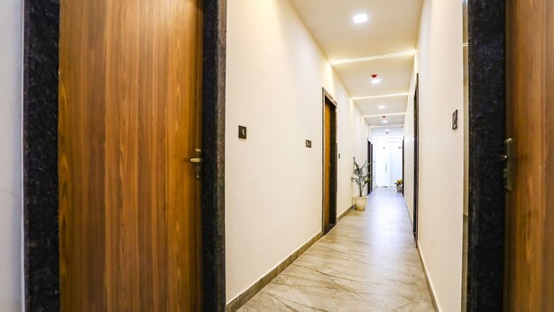 A corridor with wooden doors and marble flooring, leading to a bright room with a plant. - Fabhotel Vasudha (Mathura)