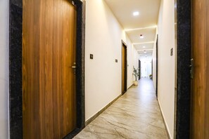 A corridor with wooden doors and marble flooring, leading to a bright room with a plant. - Fabhotel Vasudha (Mathura)