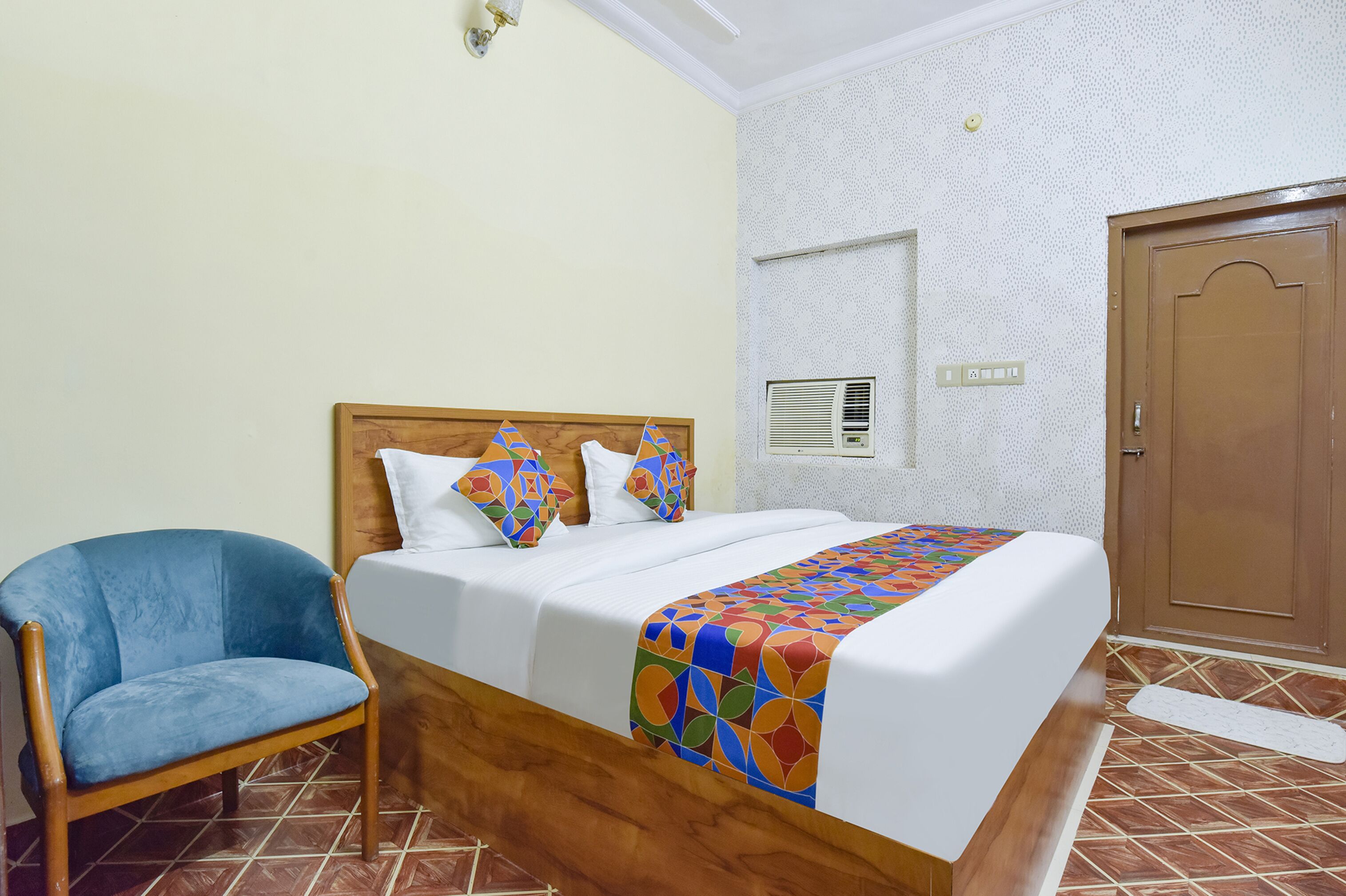 A hotel room with a bed, a blue chair, a wooden headboard, a patterned bedspread, and a tiled floor.