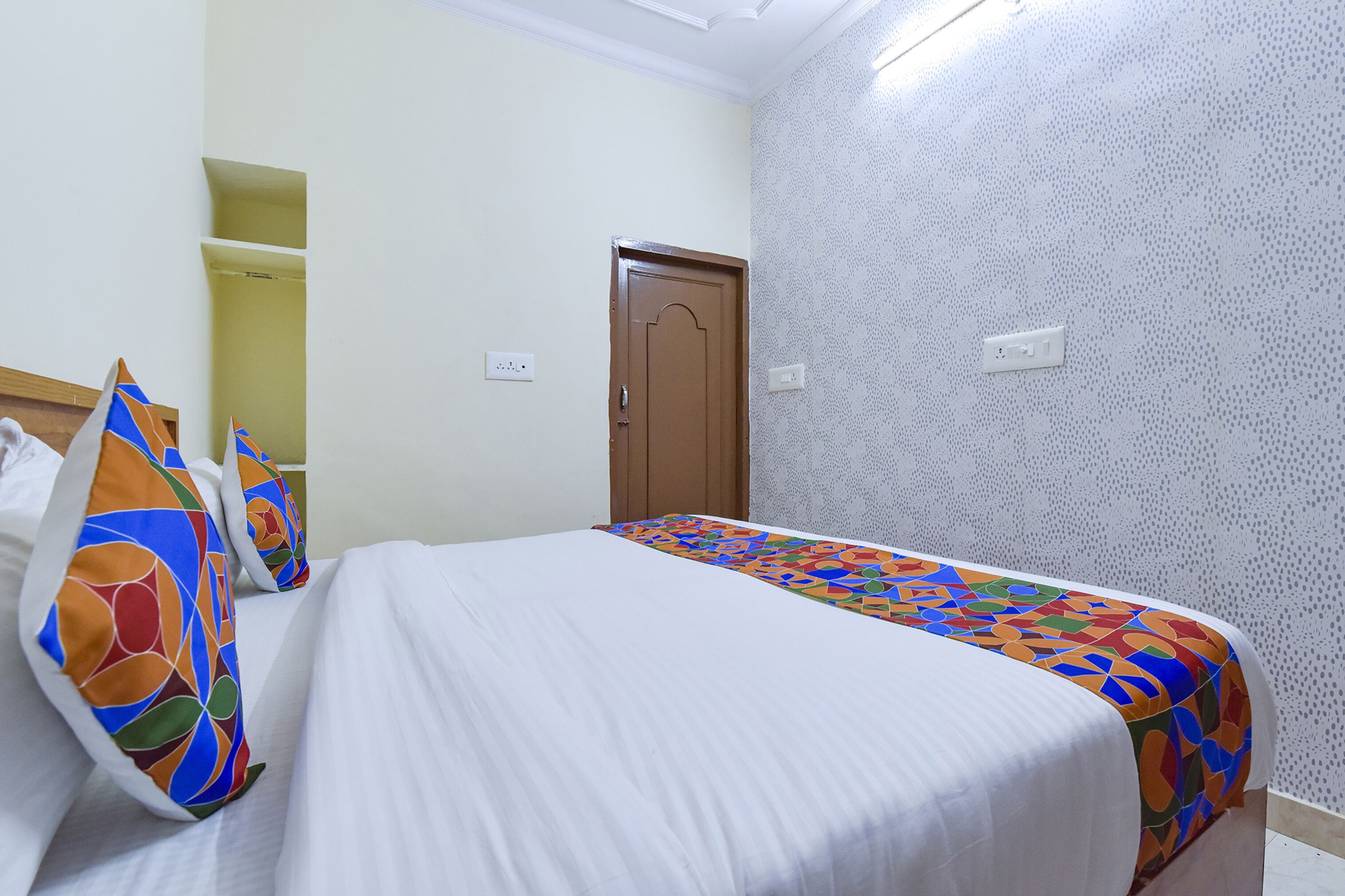 A hotel room with a bed, colorful pillows, a patterned bedspread, a wooden door, and a patterned wall.