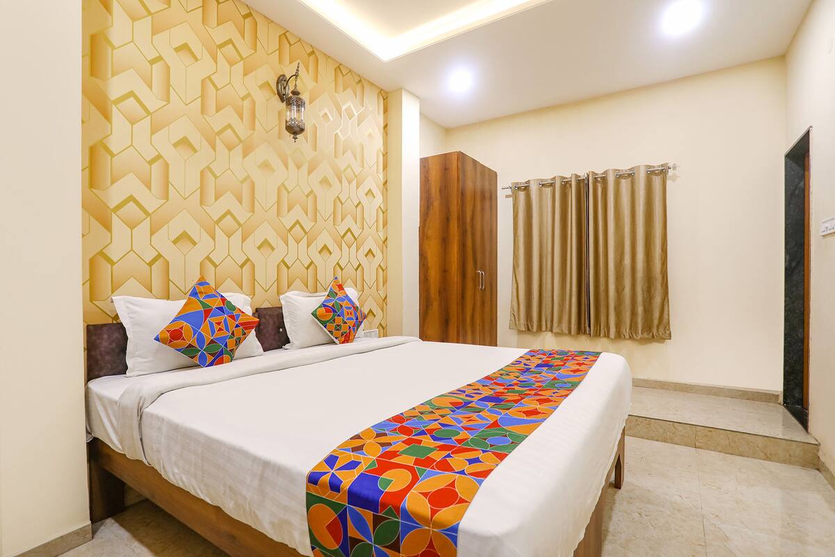 A neatly made bed with a colorful geometric bedspread, a wooden wardrobe, and a patterned wall.