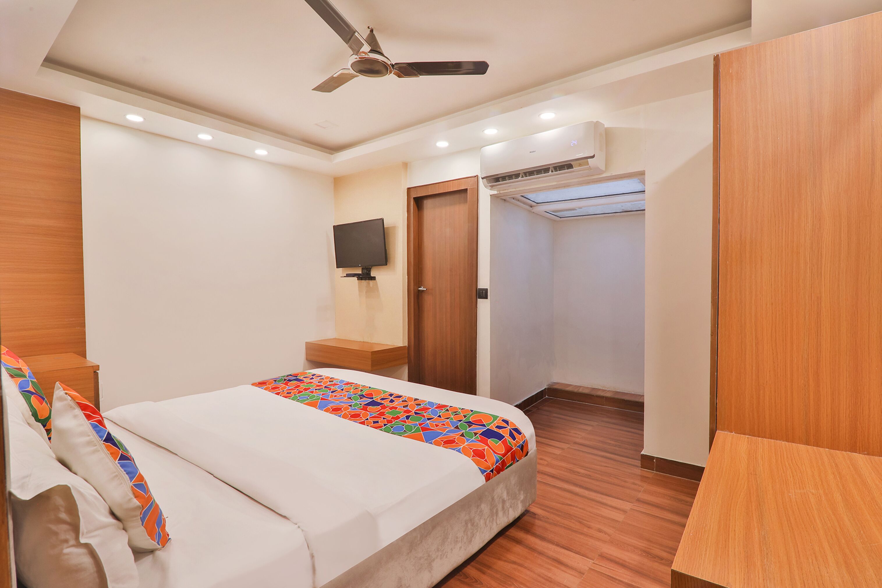 A hotel room with a bed, a ceiling fan, a TV mounted on the wall, an air conditioning unit, and a wooden wardrobe.