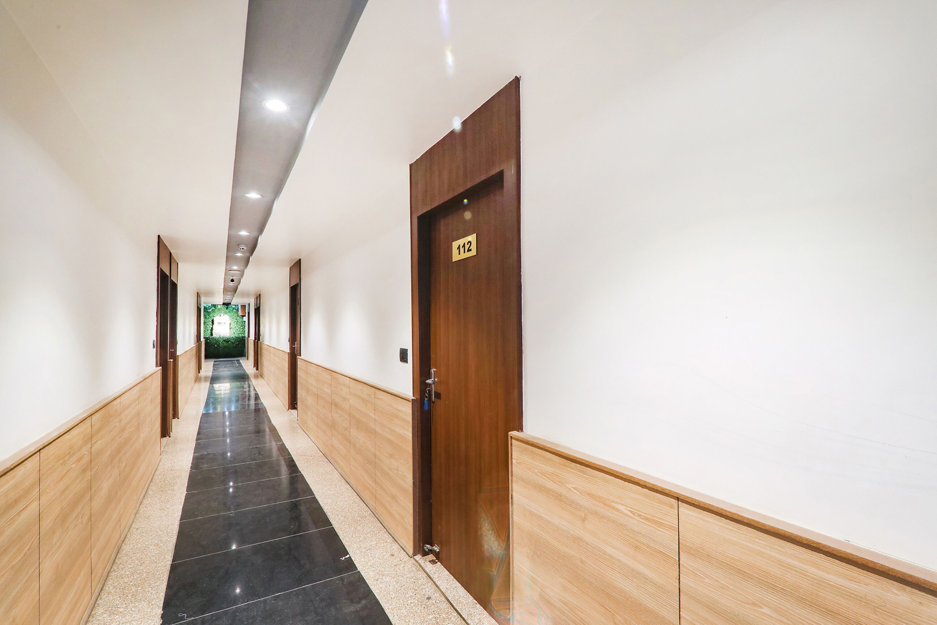 Un pasillo largo y estrecho con una puerta de madera etiquetada con el número “112” a la derecha. El pasillo tiene un piso de baldosas con un diseño negro y beige.