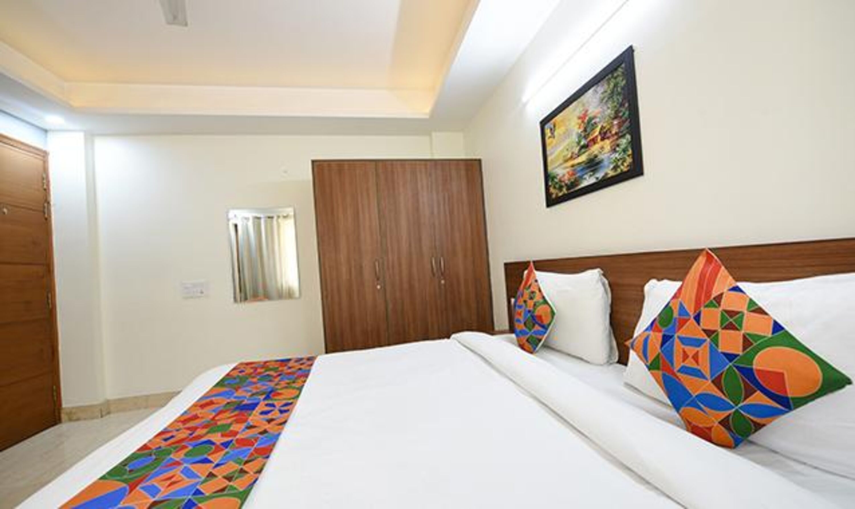 A hotel room with a large bed, a wooden wardrobe, a painting on the wall, and a window with blinds.