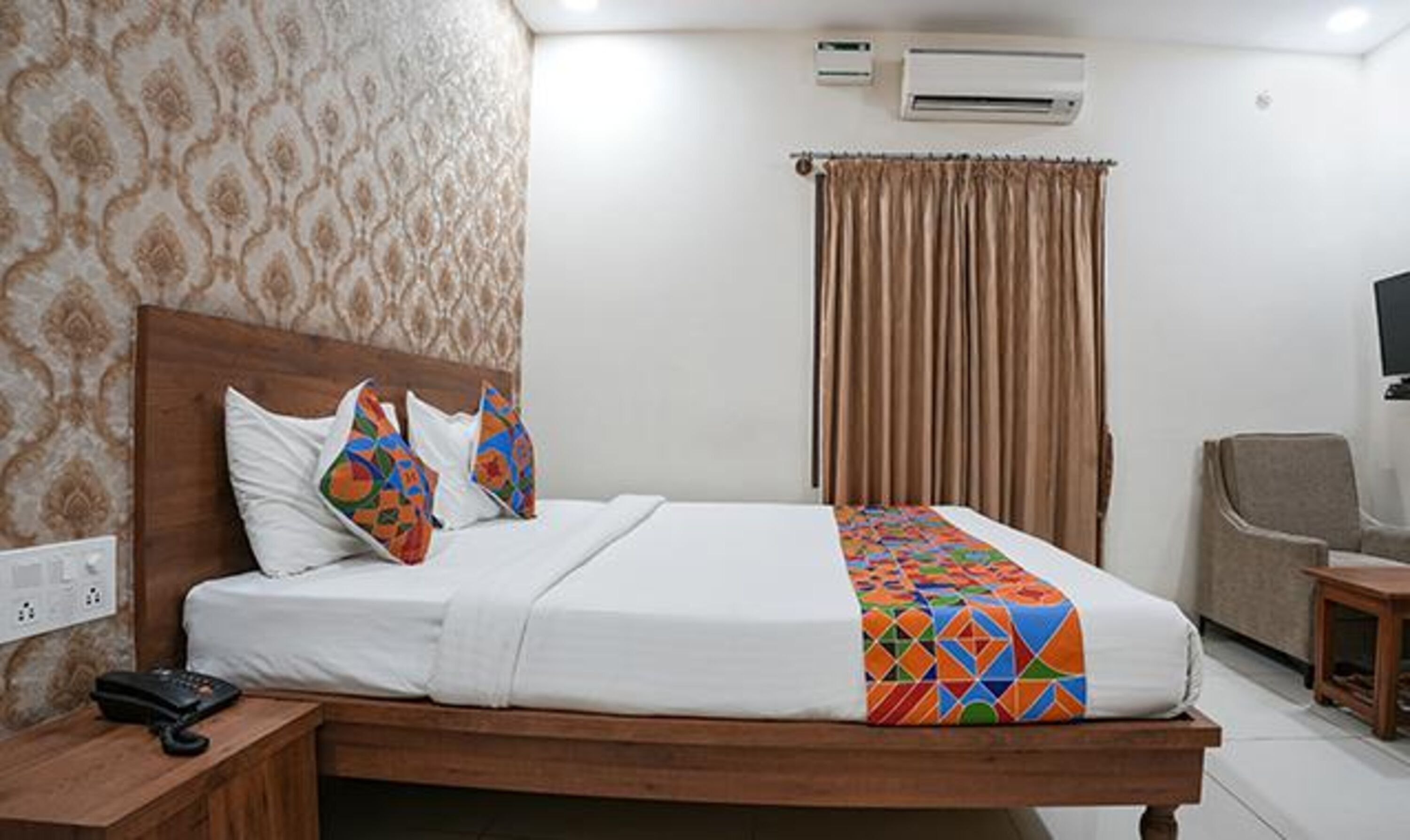 A hotel room with a wooden bed, white bedding, a colorful geometric throw blanket, a telephone, an air conditioning unit, and a television.
