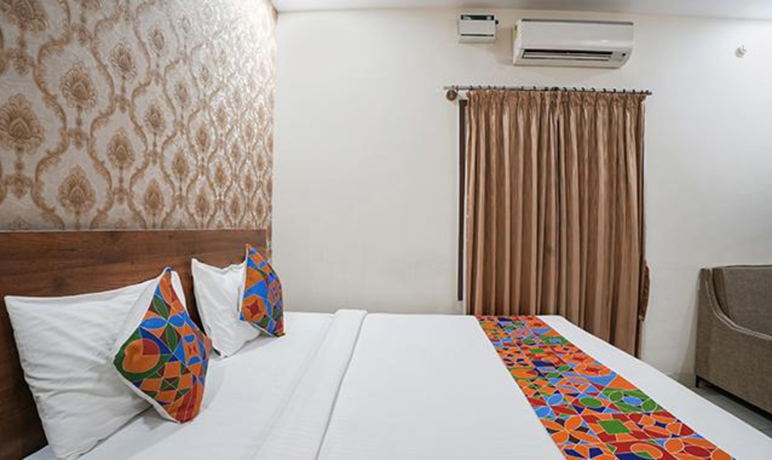 A hotel room with a bed, two pillows, a colorful bedspread, an air conditioning unit, a curtain, and a chair.
