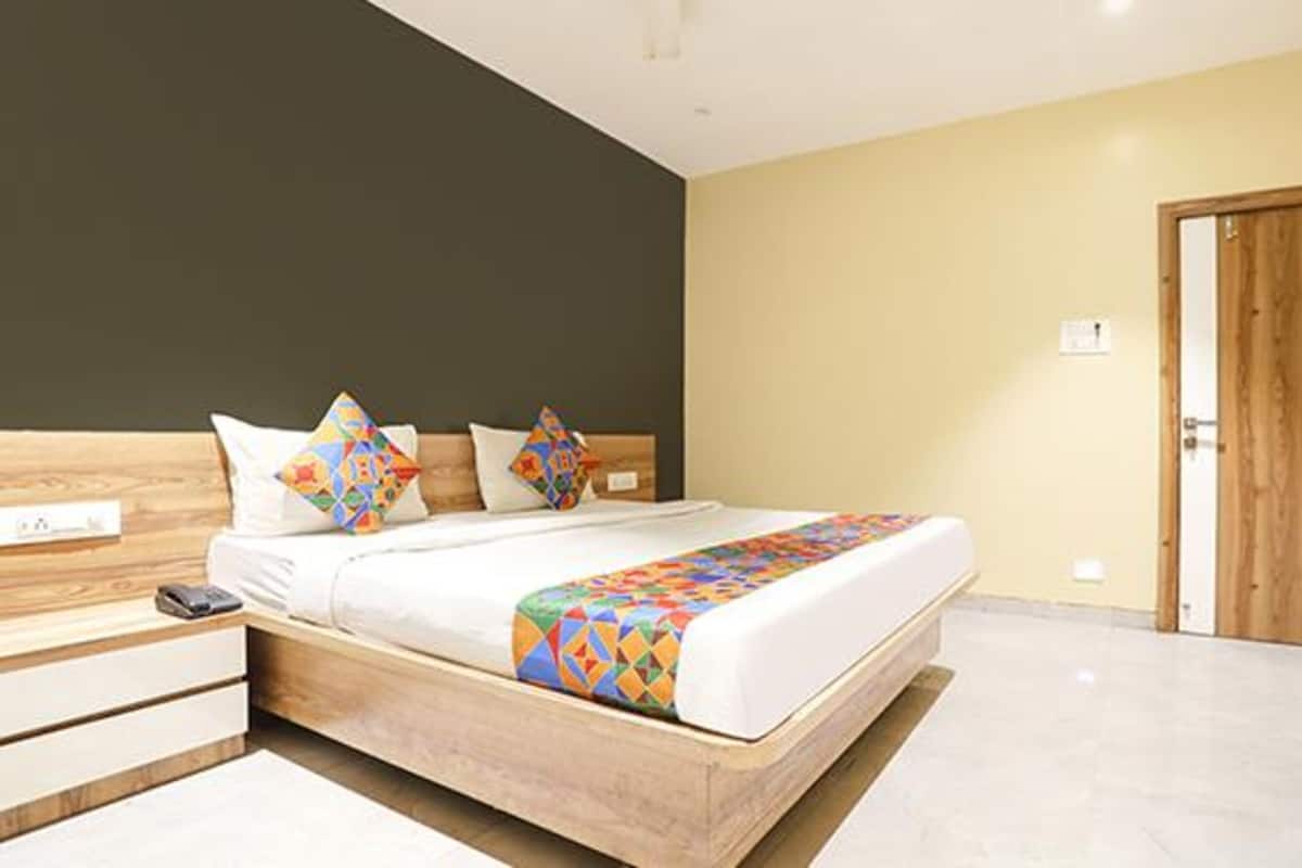 A modern bedroom with a wooden bed frame, a white mattress, and colorful geometric pillows. There is a wooden nightstand with a phone and a small lamp. The room has a beige wall and a wooden door.
