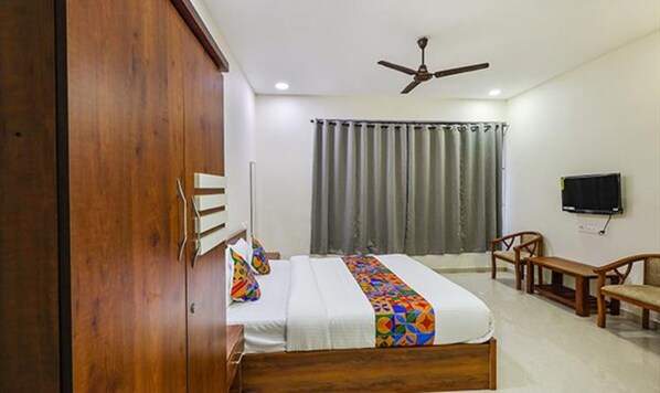 A bedroom with a wooden wardrobe, a bed with a colorful blanket, a ceiling fan, a TV, and a window with curtains. - Fabhotel De Luxe (Chennai)